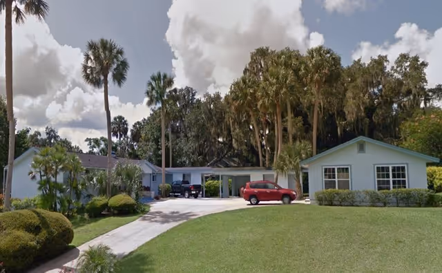 Single-story residential building with a driveway and two parked cars, surrounded by palm trees and well-maintained green lawn under a partly cloudy sky.