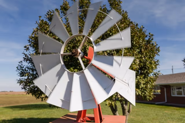 A close-up view of a white and red windmill structure with a tree and a brick building in the background under a clear blue sky.