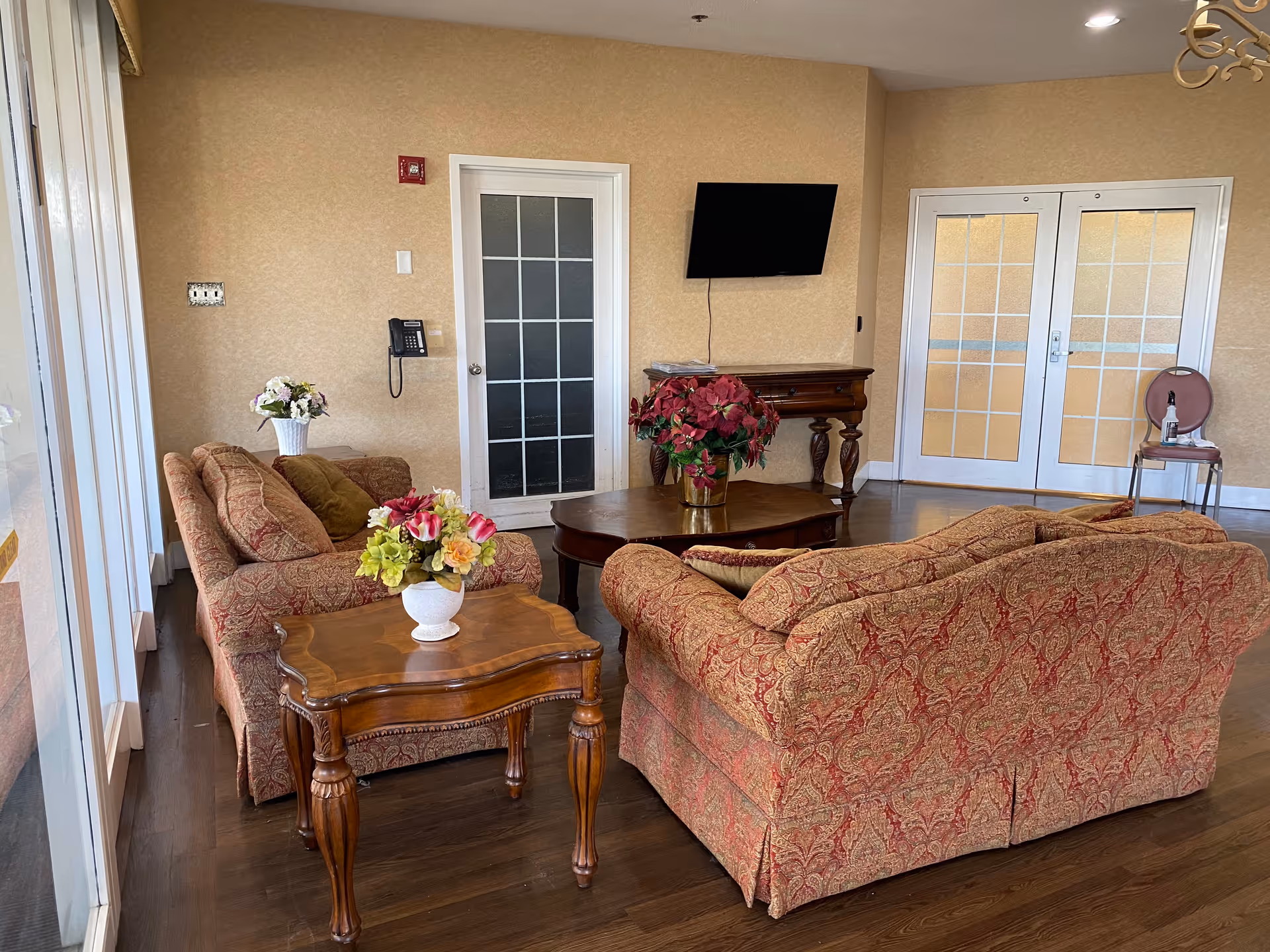 A comfortable lounge with patterned sofas and wooden tables topped with flower arrangements, a wall-mounted TV, and glass doors.