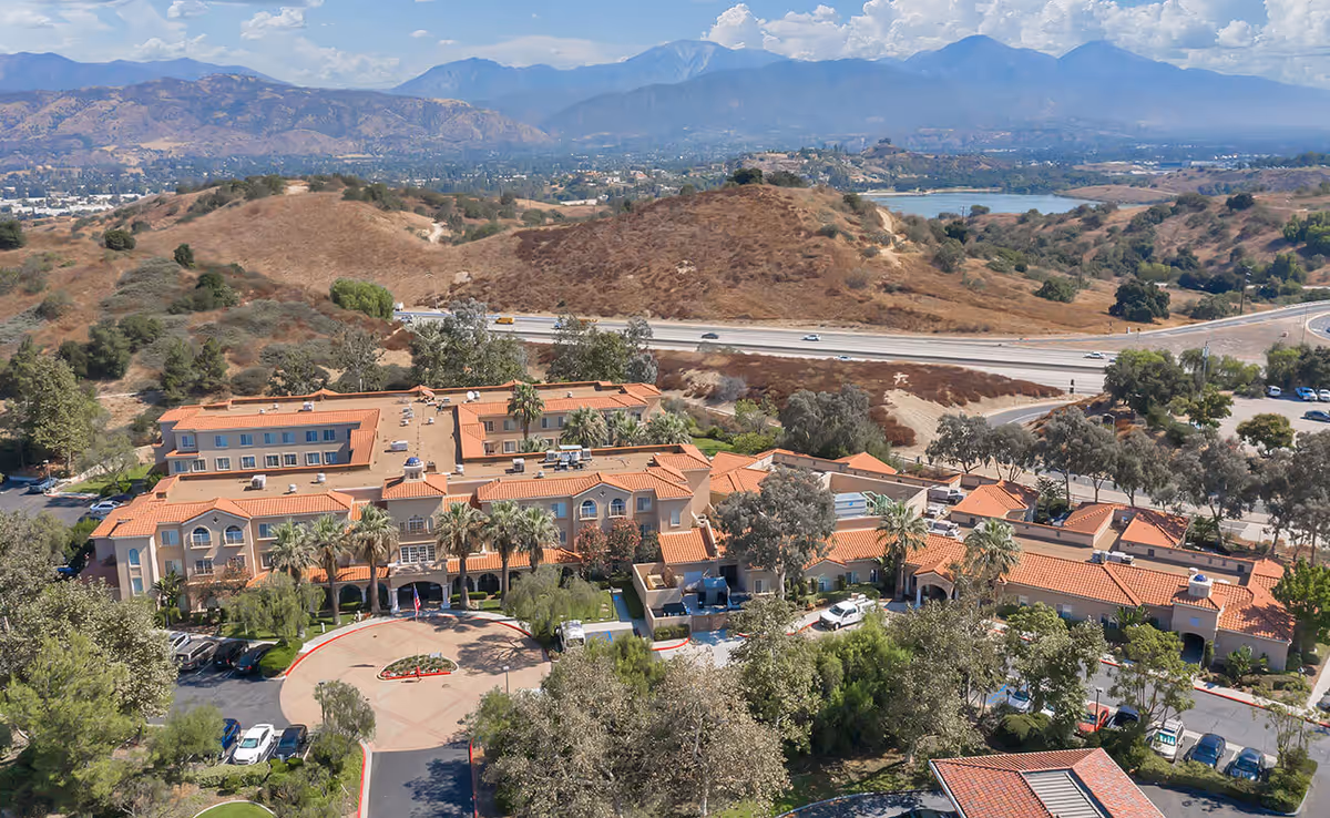 Aerial view of a senior living complex with terracotta roofs, a circular driveway and surrounding hills.