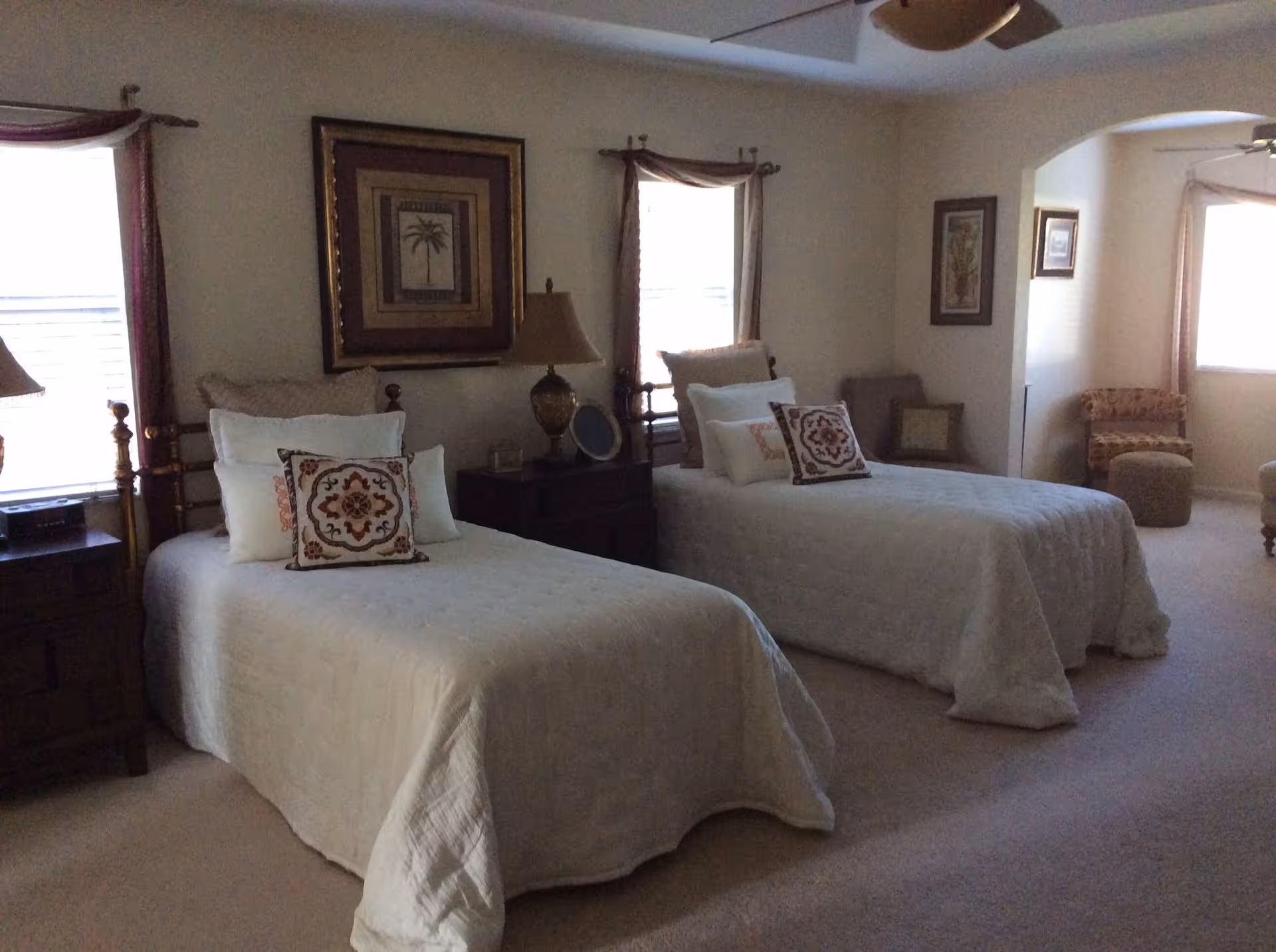 A bedroom with two twin beds, each made with white bedding and decorative pillows. Between the beds is a nightstand with a lamp and a small framed picture. The room has two windows with curtains, framed artwork on the walls, and additional seating including a chair and ottoman near an arched doorway.
