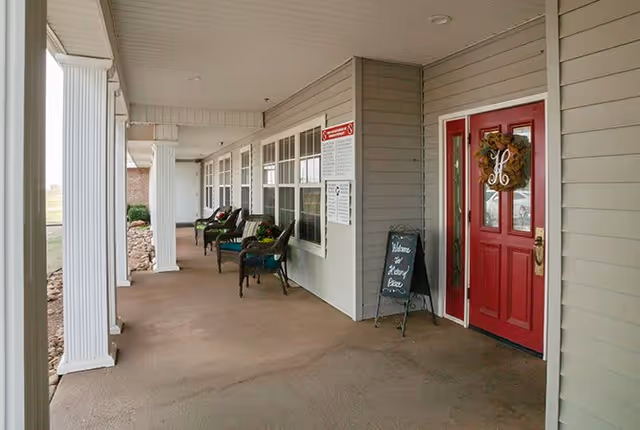 Covered porch area with white columns, several chairs with cushions, a red door decorated with a wreath, and a small chalkboard sign that says 'Welcome to Hickory Terrace'.