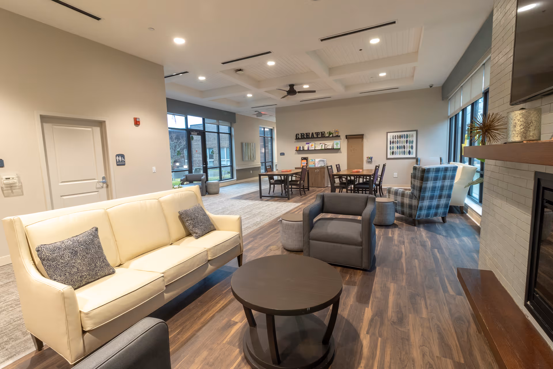 Bright communal lounge in a senior living facility with sofas, armchairs, tables, and large windows.
