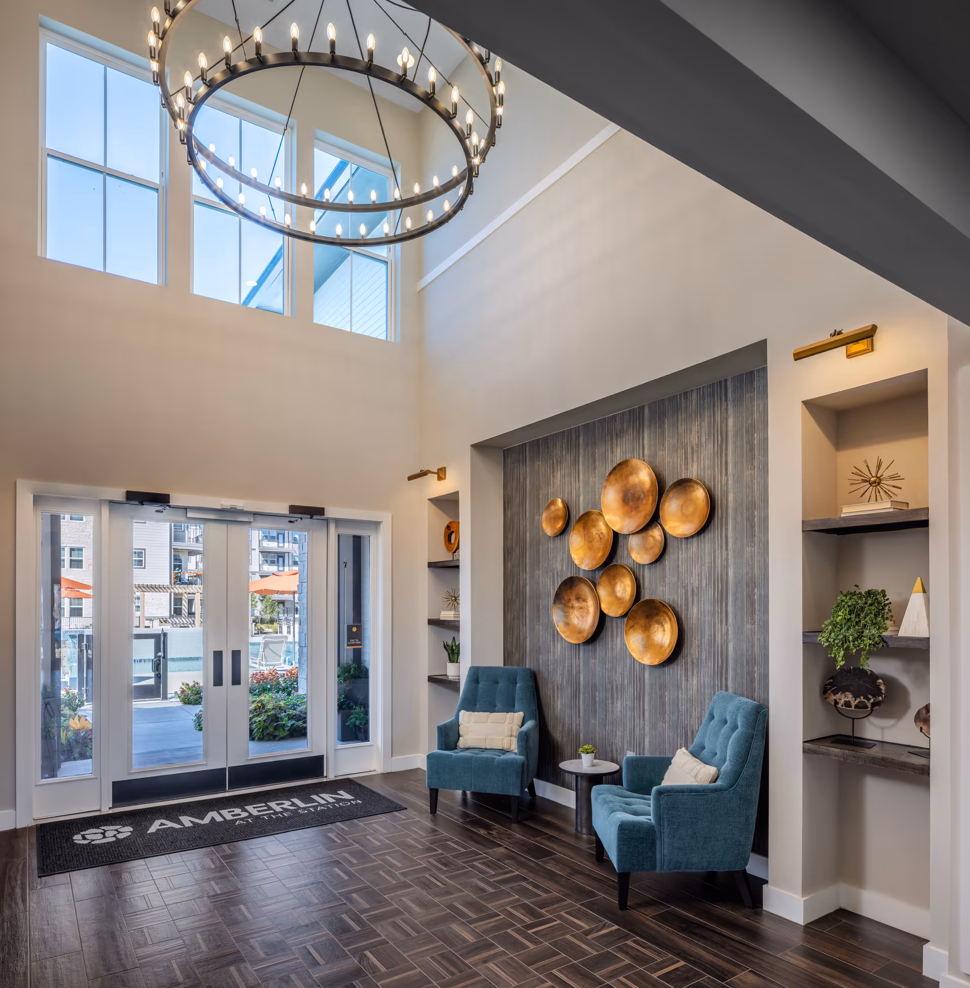 Bright and modern lobby area with high ceilings and large windows. Two teal upholstered chairs with beige pillows are placed around a small round table. The wall behind the chairs features a decorative arrangement of round, bronze-colored metal plates. Built-in shelves on either side hold decorative items and plants. Double glass doors lead outside, with a black mat in front that reads 'Amberlin at The Station'. A large circular chandelier hangs from the ceiling.