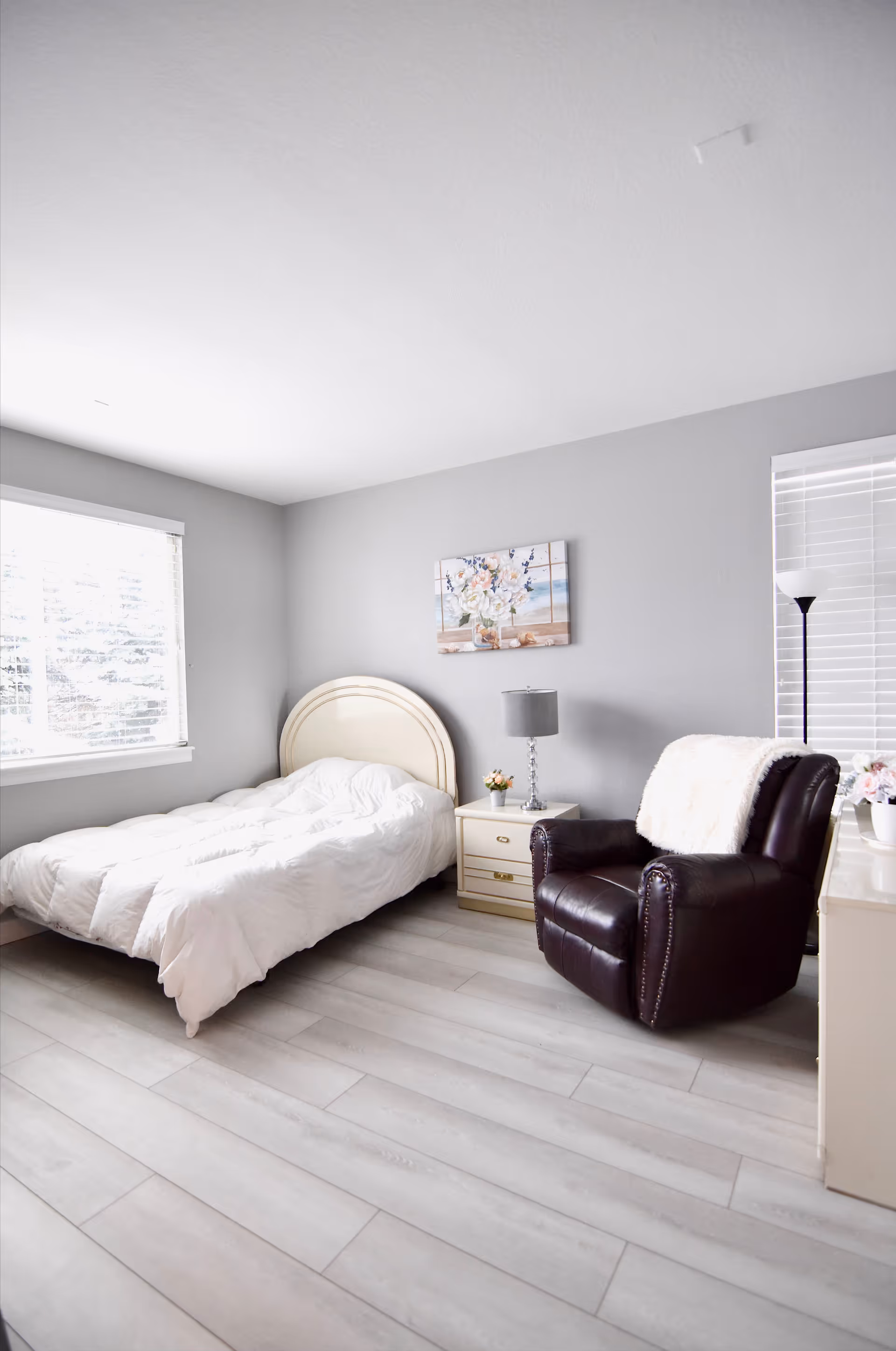 Bright, minimal bedroom with a single bed dressed in white, a nightstand and lamp, and a dark leather recliner by a window.