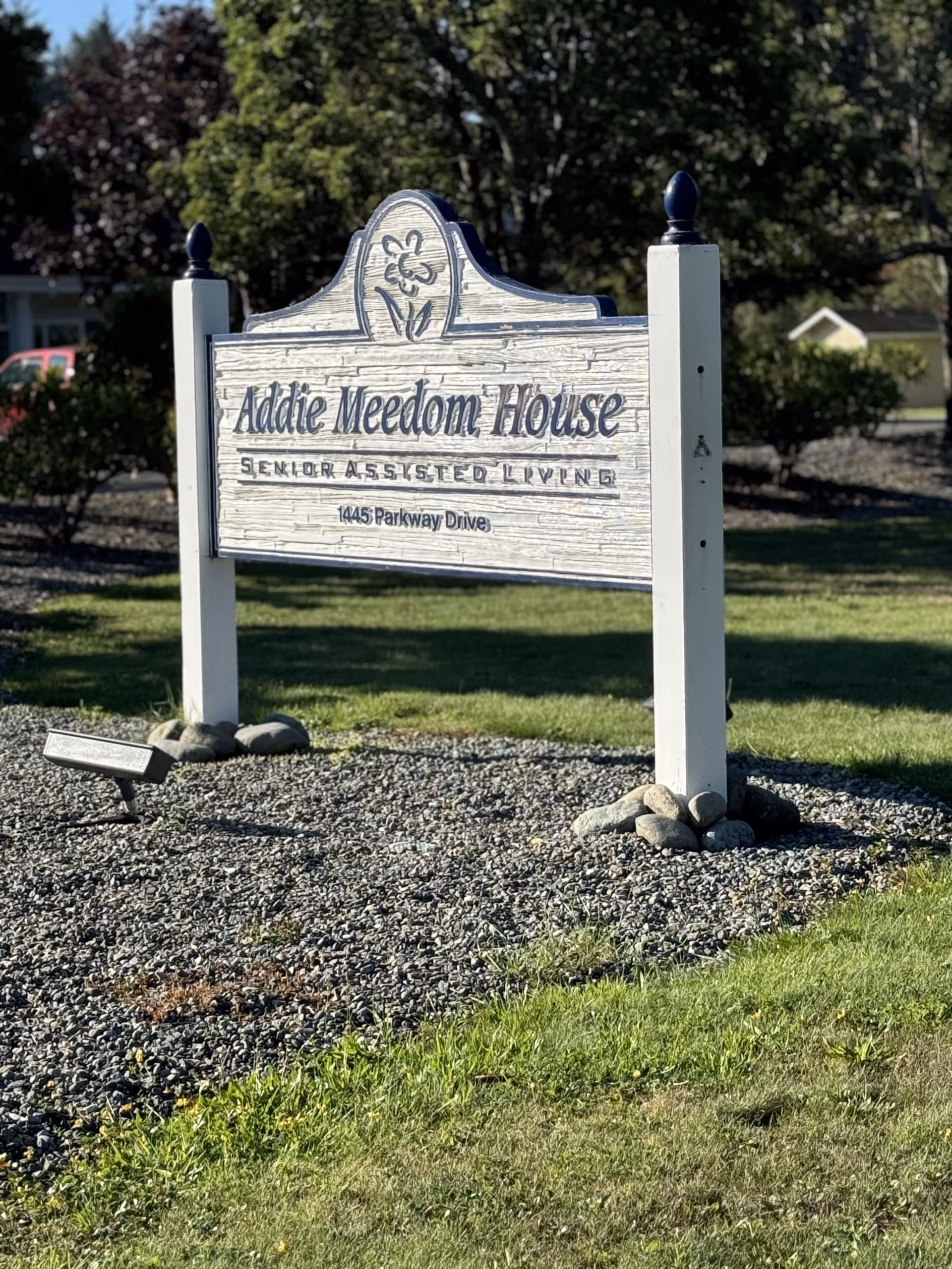 A white wooden sign with decorative top and flower design that reads 'Addie Meedom House Senior Assisted Living 1445 Parkway Drive' set on a gravel bed with grass and trees in the background.