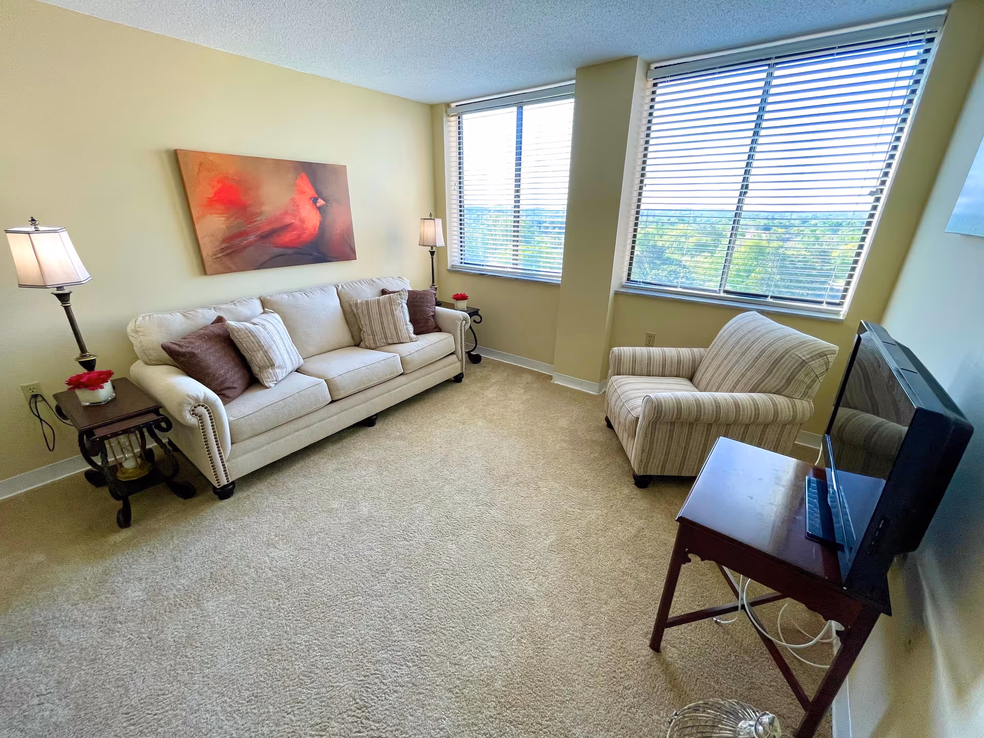 A cozy living room with a beige couch adorned with multiple pillows, a striped armchair, two floor lamps on either side of the couch, a small wooden side table, a TV on a wooden stand, and two large windows with blinds letting in natural light. A painting of a red bird hangs on the wall above the couch.