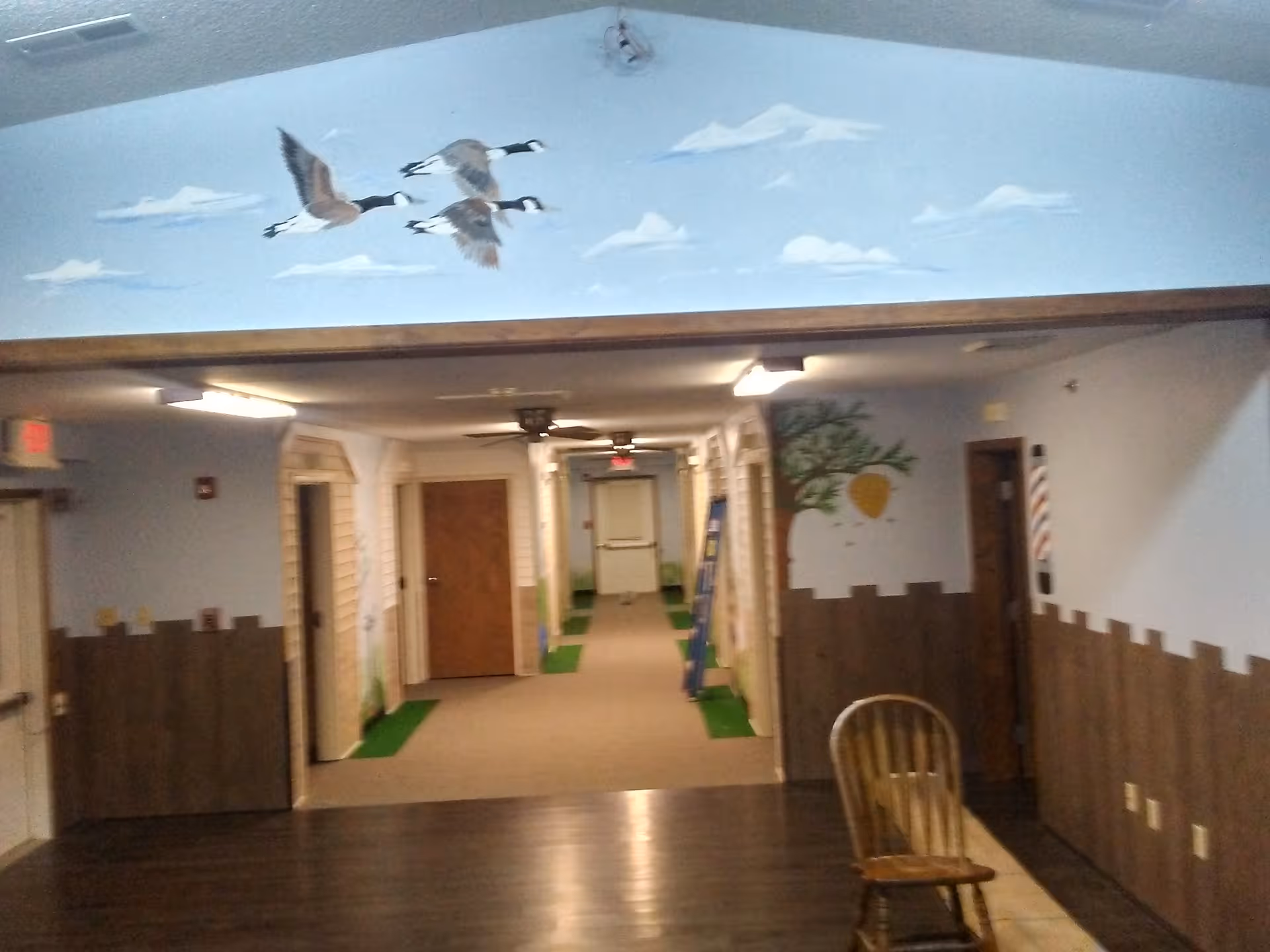 Interior hallway of a senior living facility with painted murals of flying geese and a tree, doors along the corridor and a wooden chair in the foreground.