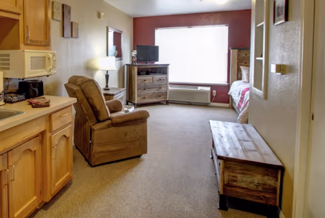 Interior view of a senior living facility room featuring a small kitchenette with wooden cabinets and a microwave on the left, a brown recliner chair, a wooden dresser with a TV on top near a large window with blinds, and a bed partially visible on the right side against a red accent wall.