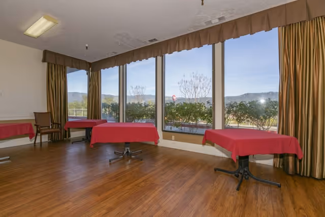 A dining area with several tables covered in red tablecloths and a few chairs. Large windows with brown curtains provide a view of greenery and distant hills outside. The room has wooden flooring and a ceiling light fixture.