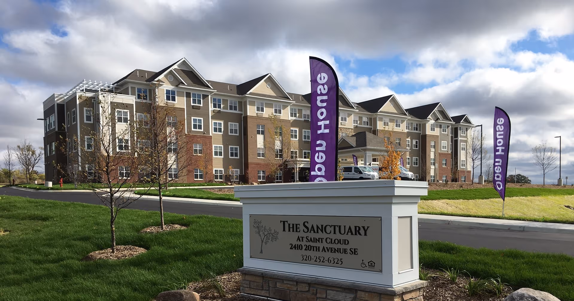 Exterior view of The Sanctuary at Saint Cloud, a multi-story senior living facility with beige, brown, and white siding. In the foreground is a white sign with the facility's name, address, and phone number. Two purple flags with white text are visible near the entrance. The sky is partly cloudy.