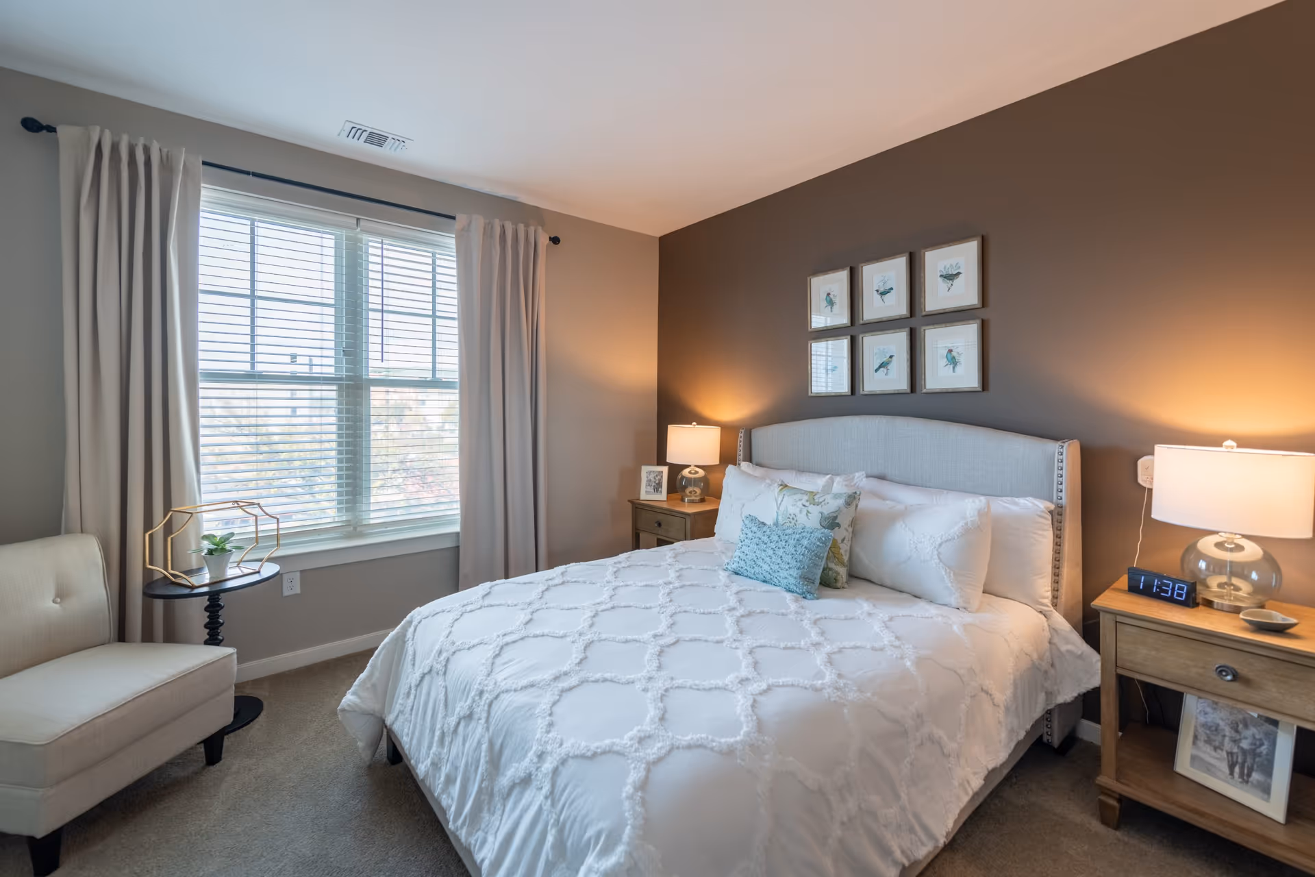 A cozy bedroom with a large bed covered in a white textured comforter and multiple pillows. The bed has a light gray upholstered headboard and is flanked by two wooden nightstands, each with a lamp. One nightstand has a digital clock showing 11:38. Above the bed, there are six framed botanical prints arranged in two rows. A window with white blinds and beige curtains lets in natural light. A beige upholstered chair and a small round black side table with a decorative plant are positioned near the window.