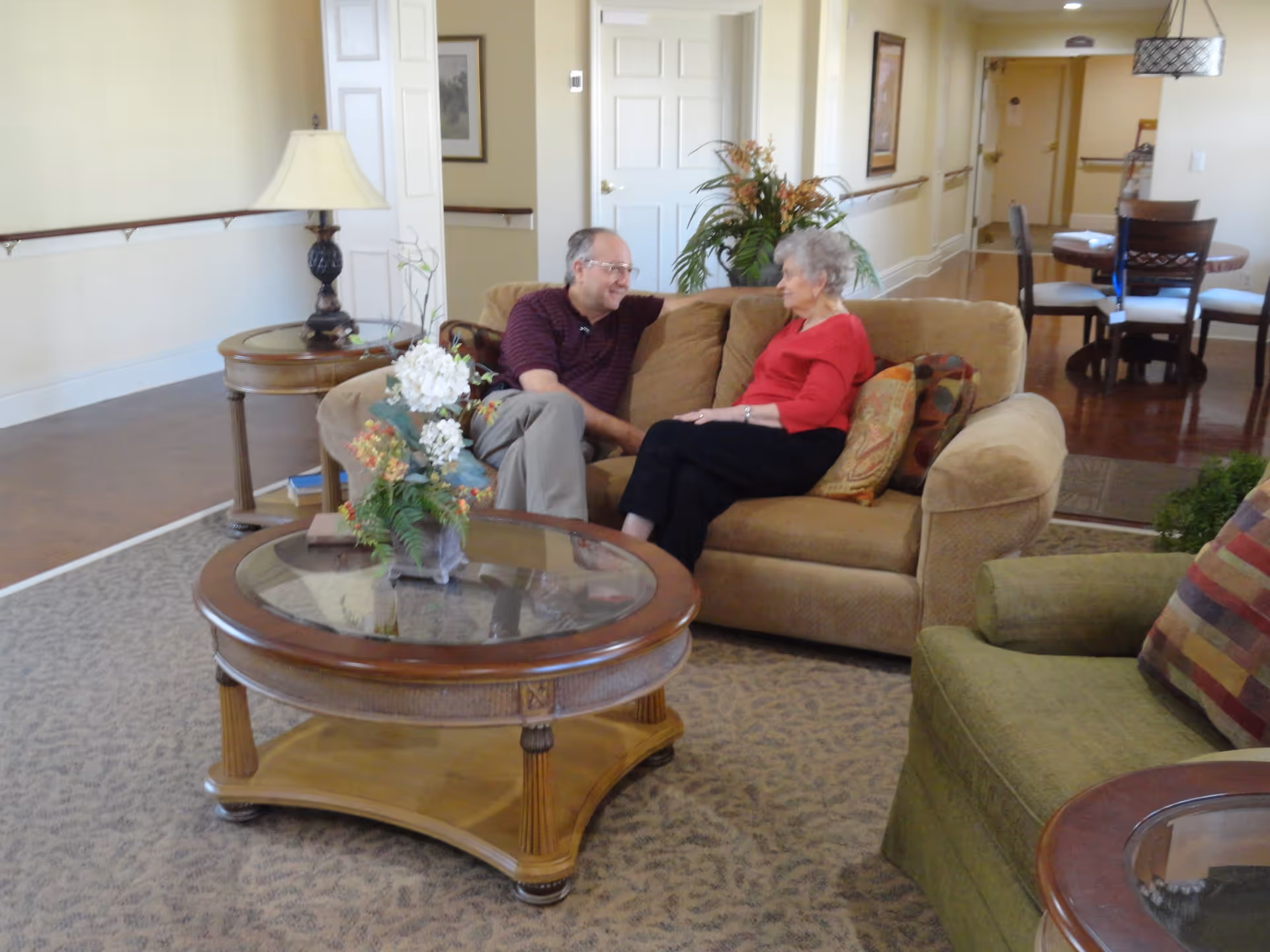 An elderly woman and a middle-aged man sitting and talking on a beige couch in a well-lit living room area with a round glass-top coffee table with a flower arrangement in front of them. There is a green armchair with a colorful pillow to the right and a round dining table with chairs in the background.