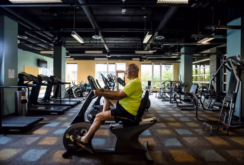 An elderly man in a bright yellow shirt and black shorts exercises on a recumbent stationary bike in a spacious gym with various exercise equipment including treadmills and weight machines. Large windows in the background let in natural light.