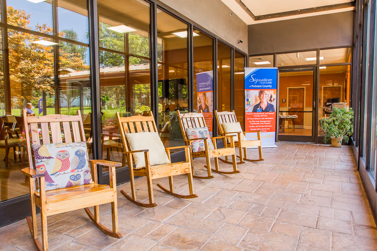 A sunlit indoor patio area with four wooden rocking chairs, each with decorative cushions. The space has large glass windows reflecting outdoor greenery and trees. A banner for Signature HealthCARE of Putnam County is visible near the entrance door, along with a potted plant on the right side.