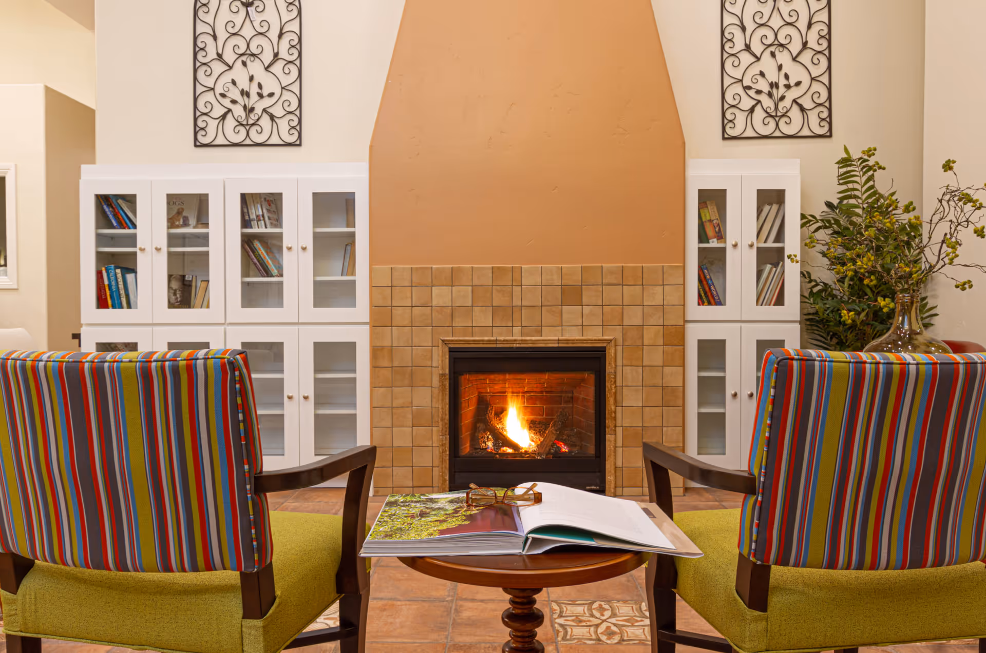 A cozy sitting area with two colorful striped armchairs facing a lit fireplace with a tiled surround. Between the chairs is a small round wooden table holding an open book and a pair of glasses. Behind the fireplace are white cabinets filled with books and decorative wrought iron wall art above.
