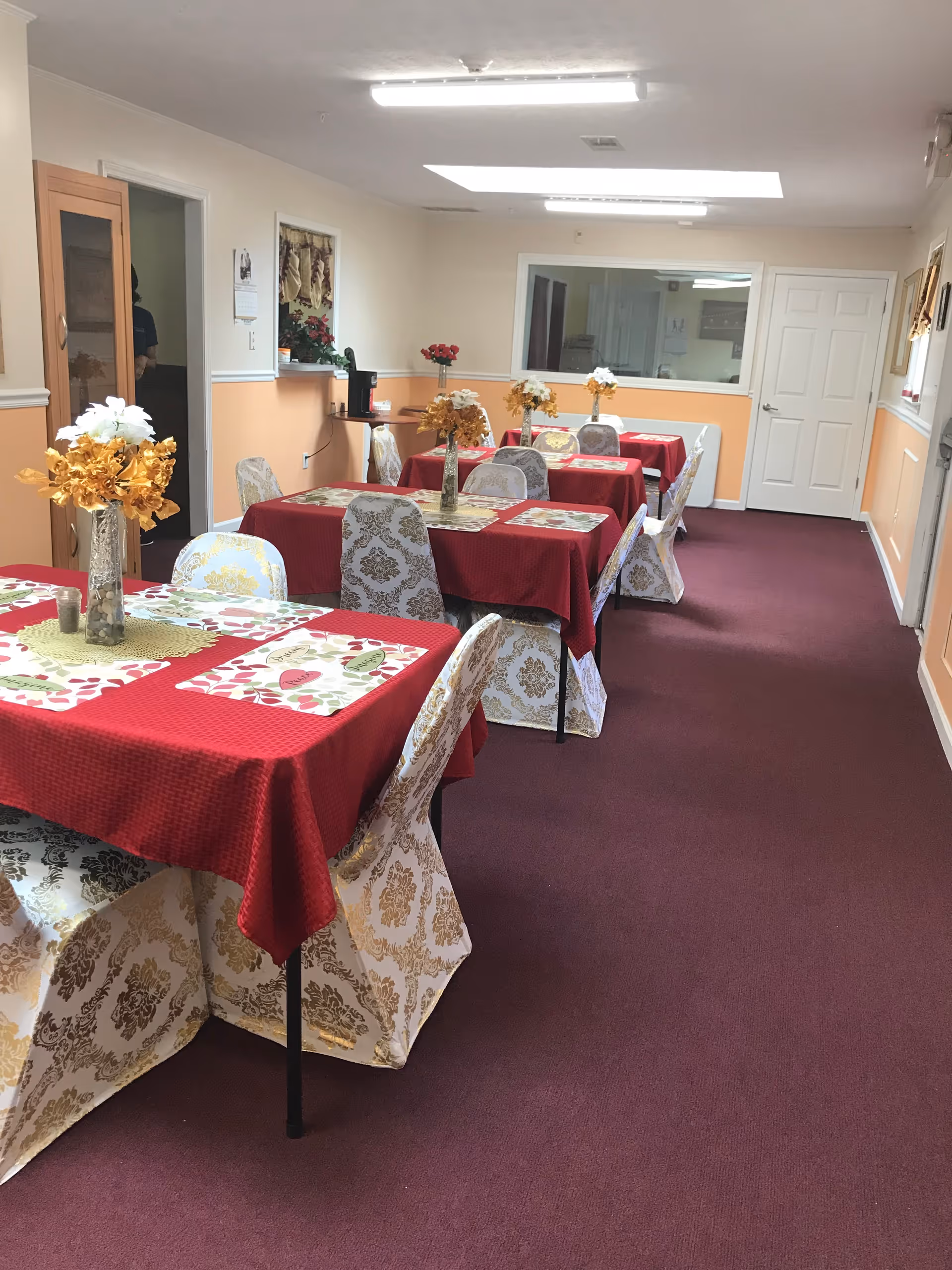 A dining room with several tables covered in red tablecloths and floral-patterned chair covers. Each table has a vase with artificial flowers as a centerpiece. The room has peach and white walls, a maroon carpet, and fluorescent ceiling lights. There is a door and a large window on the far wall, and a small counter area with a coffee maker on the left side.