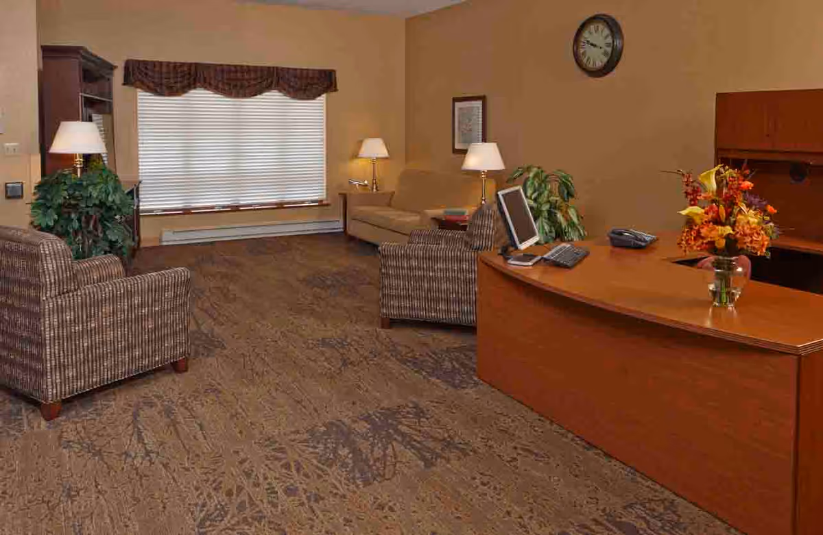 A cozy office or reception area with a wooden desk featuring a computer, phone, and a vase of flowers. The room has two patterned armchairs, a beige sofa, two table lamps, a large window with blinds and a valance, a wall clock, framed artwork, and a potted plant. The floor is carpeted with a patterned design.