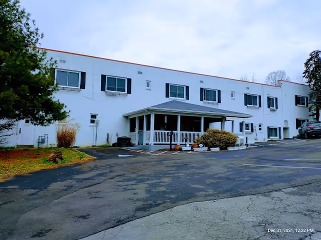 Exterior view of a two-story white building with multiple windows and a covered porch entrance. There are some bushes and trees around the building, and a paved parking area in front. The sky is cloudy.