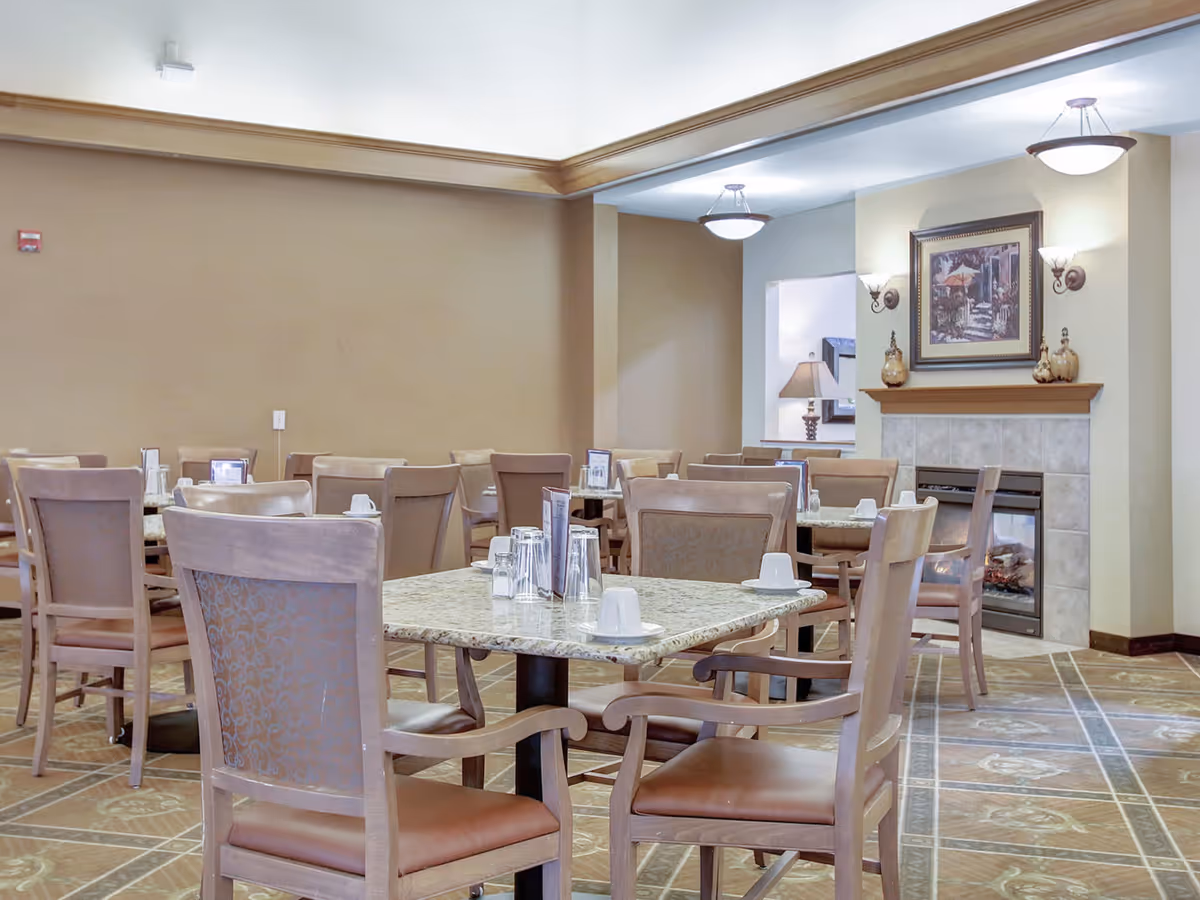 A dining room in a senior living facility with multiple tables and chairs arranged neatly. Each table has cups, glasses, and menus placed on it. The room features a fireplace with a framed painting above it, wall sconces, and ceiling lights providing warm illumination. The walls are painted in neutral tones and the floor has a patterned carpet.