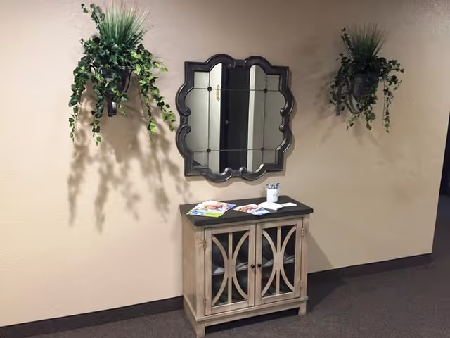 A beige wall with two hanging green leafy plants on either side of a decorative mirror with a scalloped frame. Below the mirror is a small wooden cabinet with glass doors, on top of which are some brochures and a cup holding pens. The floor is carpeted in a dark color.