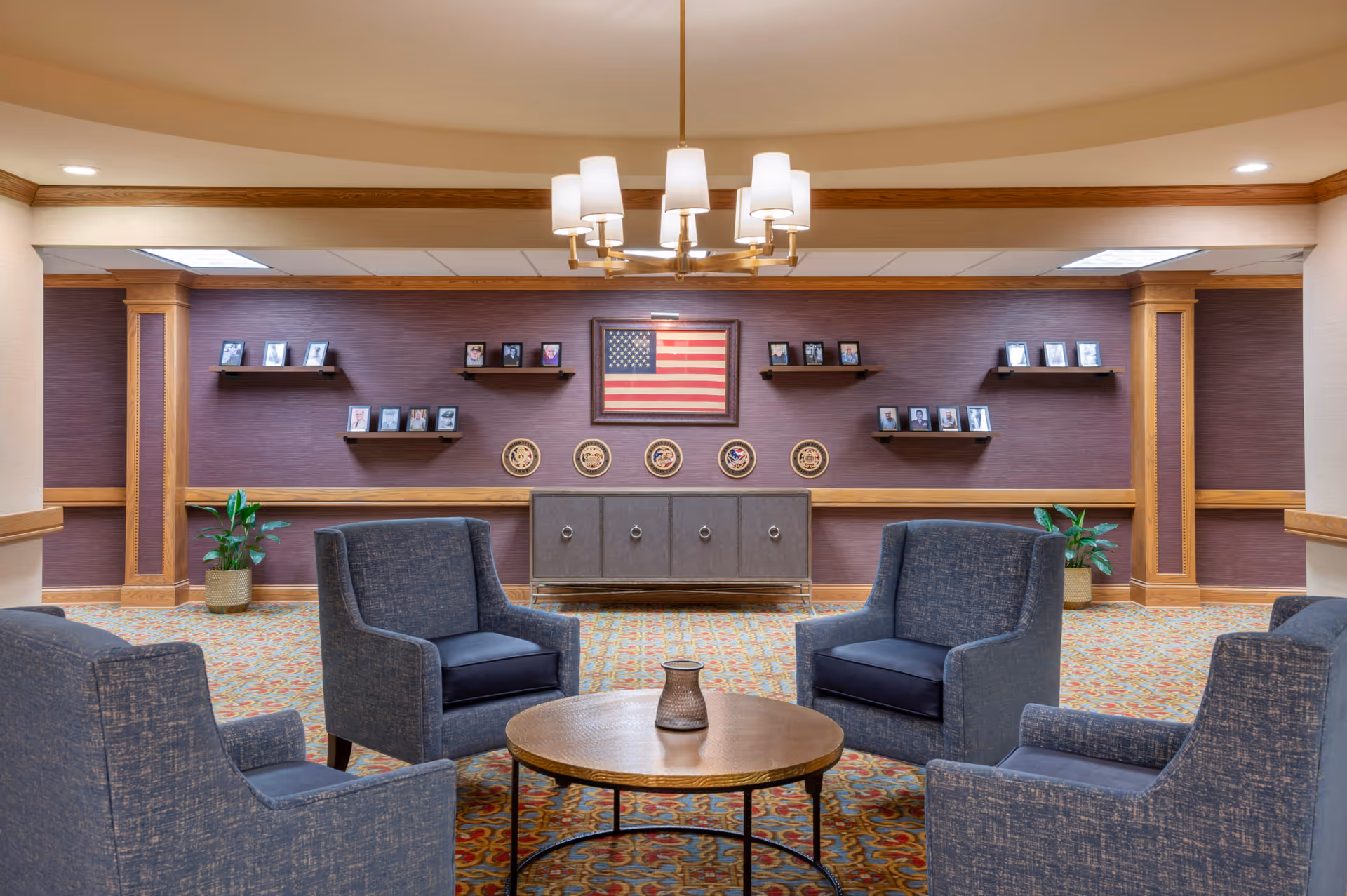 A comfortable lounge with four upholstered chairs around a round coffee table facing a wall decorated with an American flag, framed photos, and commemorative plaques.