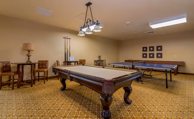 A recreational room with a pool table and a ping pong table. The room has patterned carpet flooring, beige walls, and a ceiling light fixture above the pool table. There are two chairs and a small round table with a lamp in the corner, and a rack with pool cues against the wall.