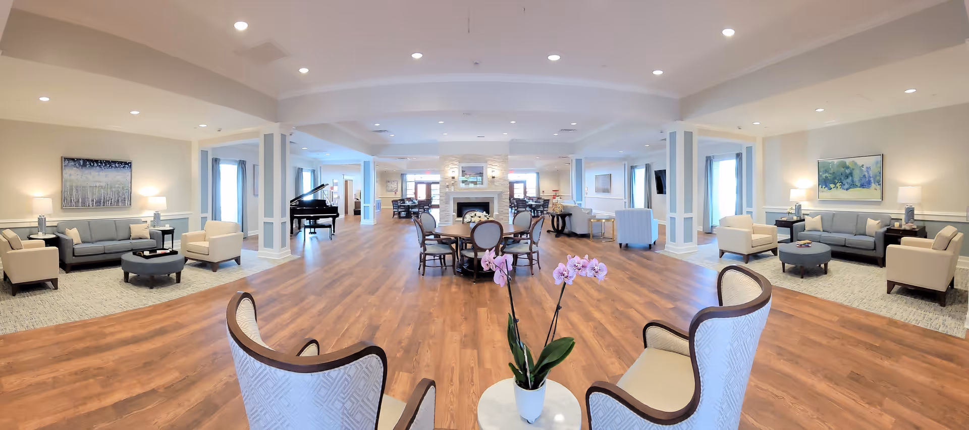 A spacious and well-lit assisted living common area with wooden flooring, multiple seating arrangements including sofas, armchairs, and round tables. A grand piano is visible on the left side, and a stone fireplace with a mirror above it is centered in the background. The room is decorated with paintings on the walls and has large windows with curtains allowing natural light to enter.