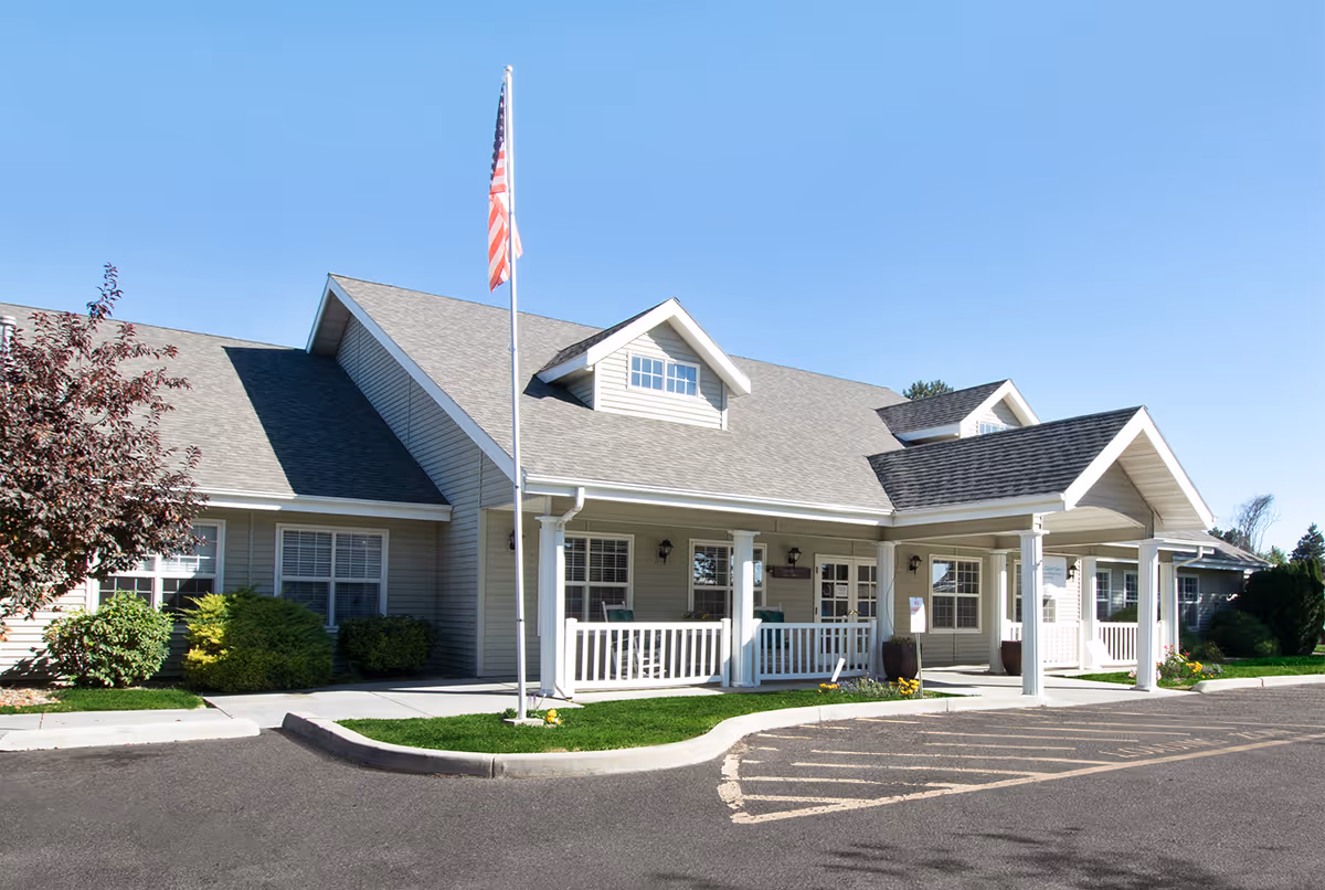 Exterior front view of a single-story senior living facility building with a covered entrance, white railings, and an American flag on a flagpole in front. The building has beige siding, multiple windows, and a gray shingled roof under a clear blue sky.
