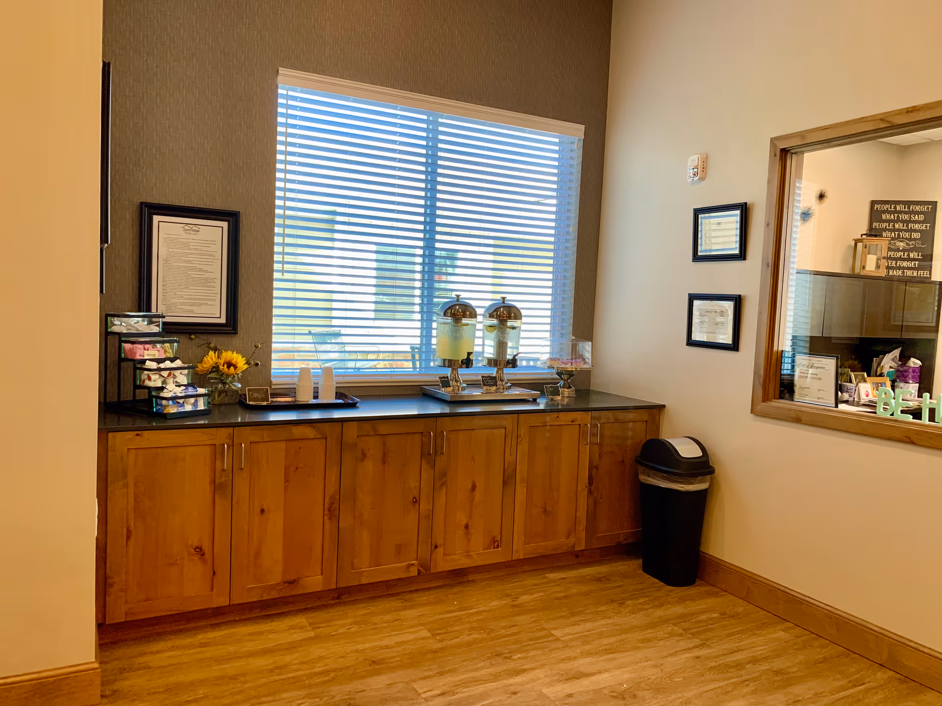 A refreshment area with wooden cabinets beneath a countertop holding two beverage dispensers, disposable cups, a small display of snacks, and a vase with sunflowers. A large window with blinds is behind the counter, and a trash can is placed next to the wall. There is a window on the right side showing an office space with framed certificates and motivational signs.
