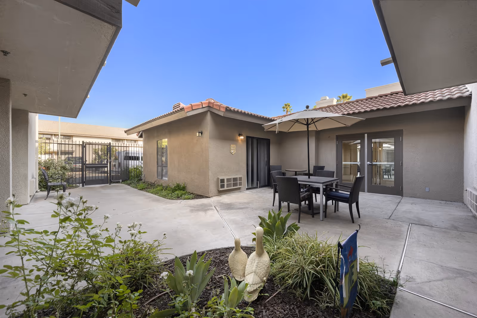 Outdoor courtyard area at Crescendo Senior Living featuring a concrete patio with a table, chairs, and an umbrella. There are plants and decorative statues in a garden bed in the foreground, and a gated entrance is visible in the background under a clear blue sky.