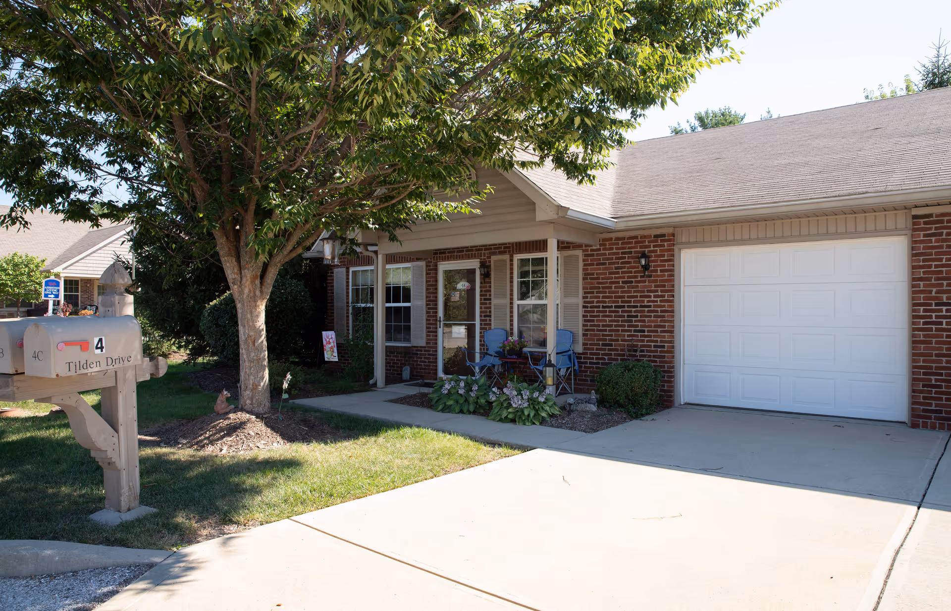 Exterior view of a single-story brick assisted living facility with a white garage door, a small porch with two blue chairs and potted plants, a tree providing shade, and a mailbox labeled '4 Tilden Drive' in the foreground.