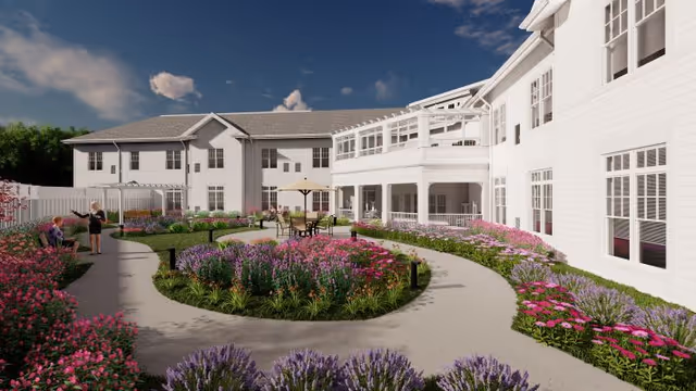 Courtyard garden with curved walking paths, flower beds, and seating in front of a white two-story senior living building.