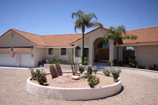 Single-story building with a red tile roof and beige exterior walls, featuring an arched entrance with two palm trees in front and a circular landscaped area with desert plants and rocks. An American flag is mounted near the garage on the left side.