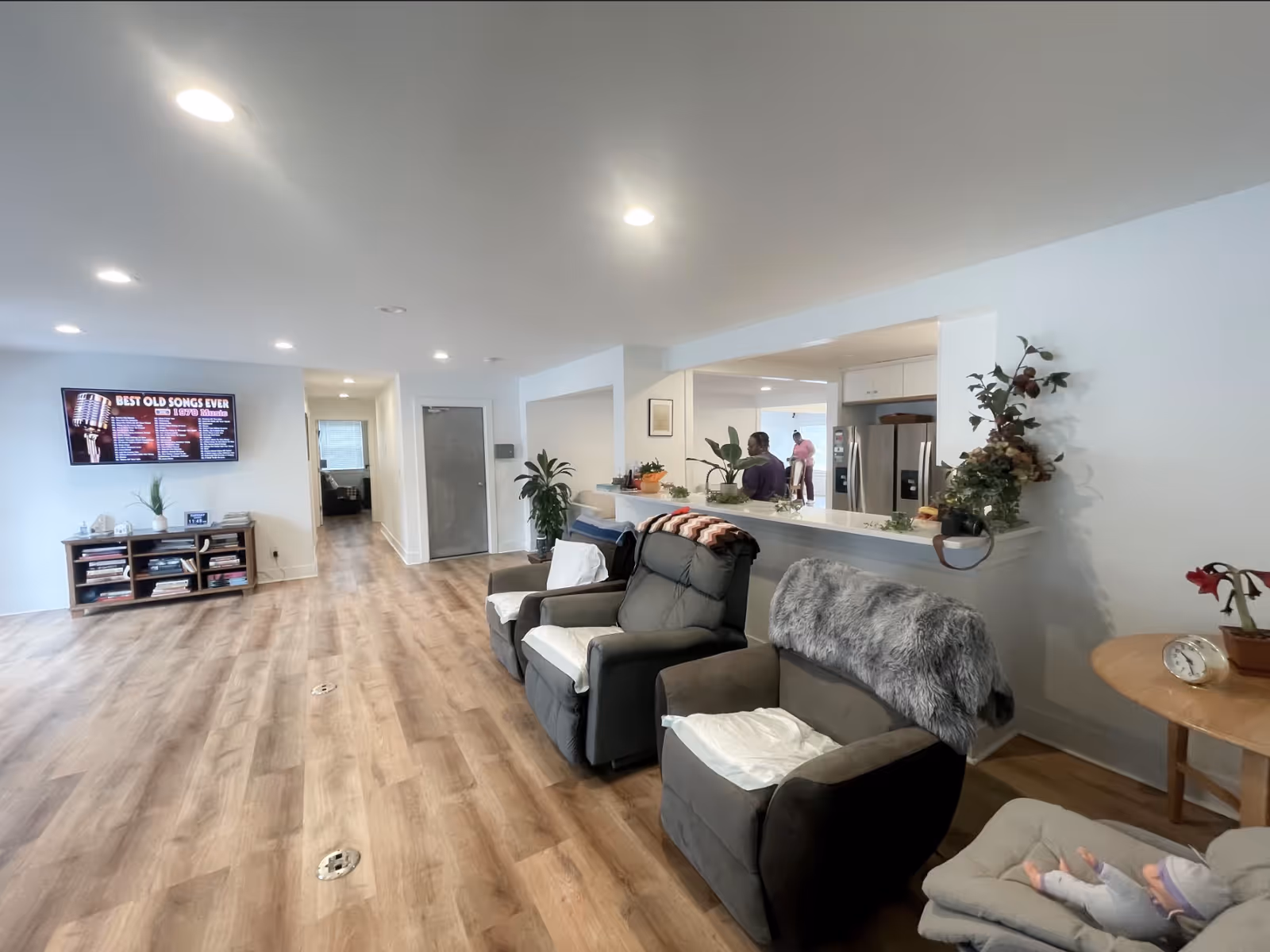 Spacious senior living common area with reclining chairs facing a wall-mounted TV, an open kitchen counter, and wood-look flooring.