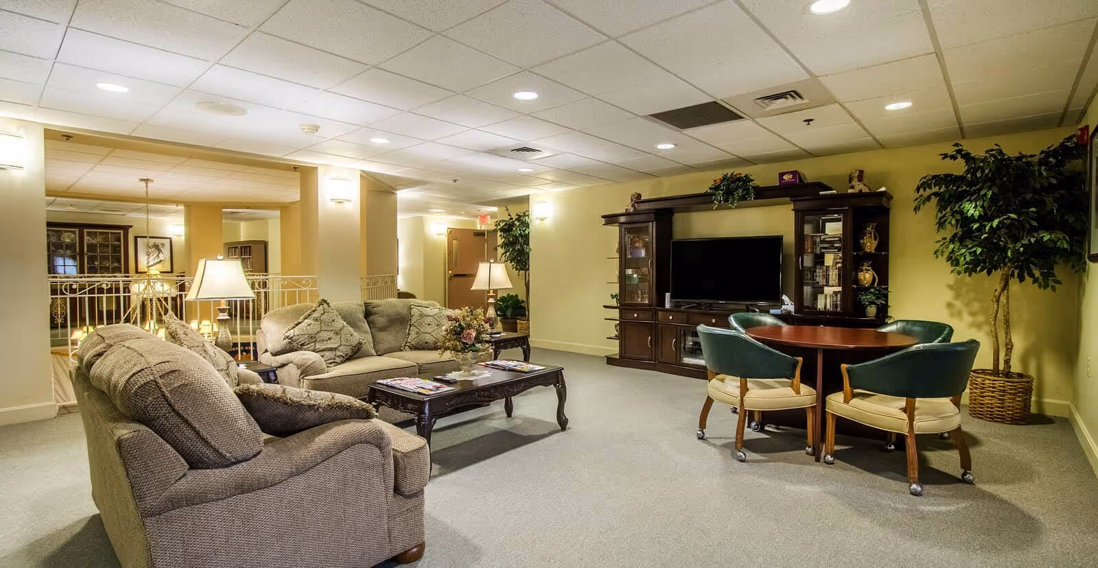 A cozy senior living community lounge area with two beige sofas, a wooden coffee table with magazines, a wooden entertainment center with a flat-screen TV, a round table with four green cushioned chairs, and a large potted plant in the corner. The room is softly lit with ceiling lights and wall sconces.