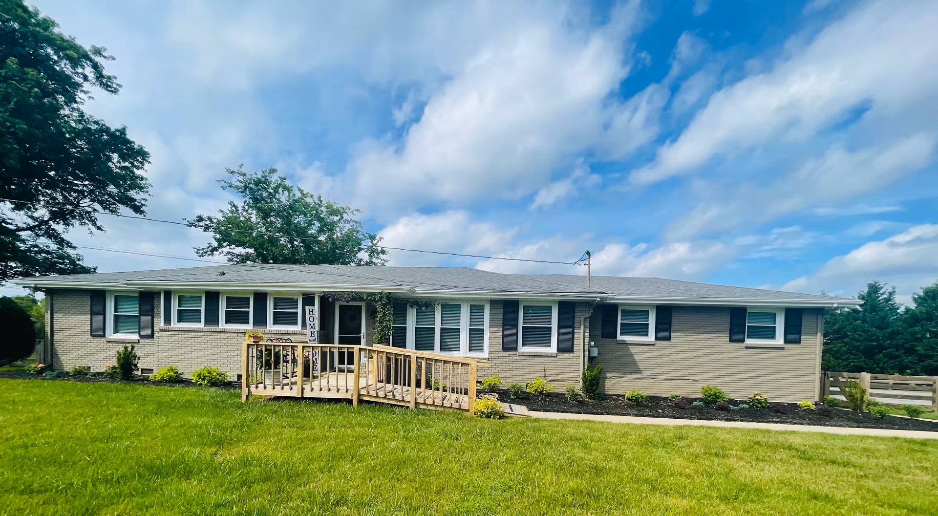 Single-story ranch-style home with a front ramp, small porch, and a manicured lawn under a partly cloudy sky.