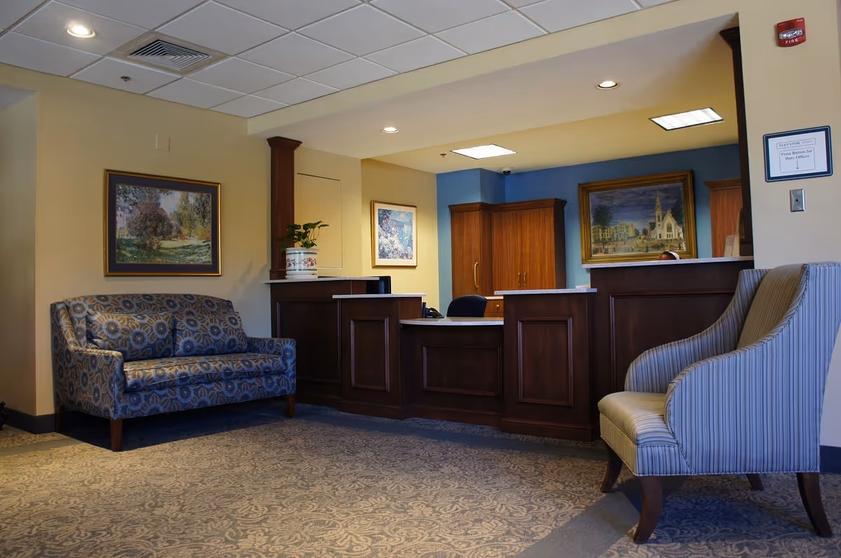 Reception lobby with a wooden front desk, patterned sofa and armchair, and framed artwork on the walls.