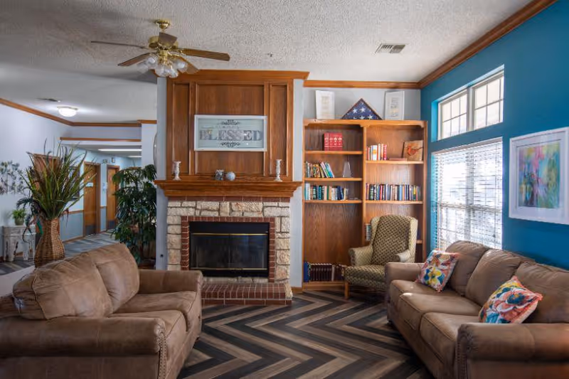 A cozy senior living common area with two brown sofas, a green armchair, a wooden bookshelf filled with books, a brick fireplace with a wooden mantle, and large windows letting in natural light. The walls are painted white and teal, and there is a ceiling fan above.