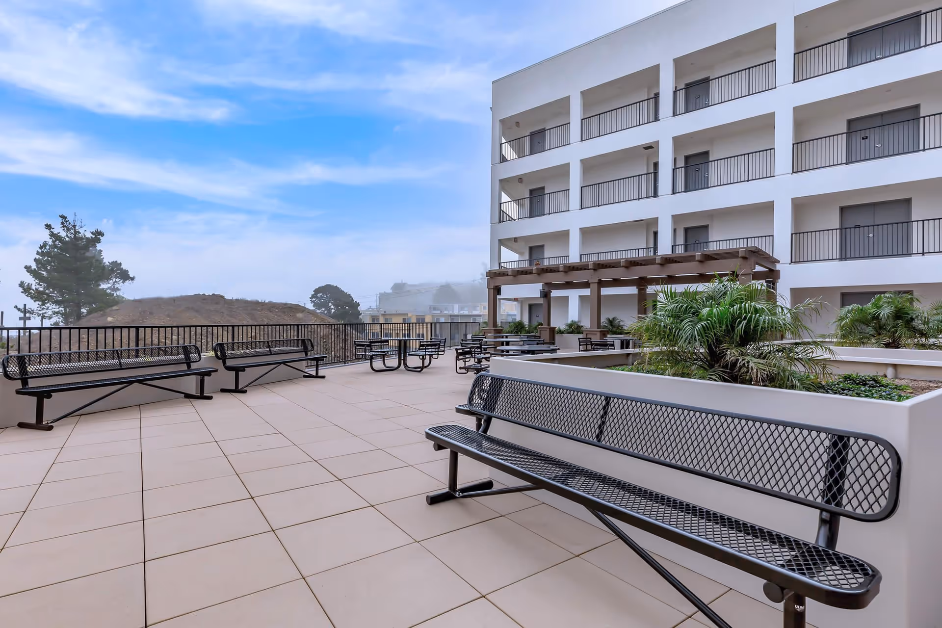 Outdoor patio area at Brunswick Street Apartments featuring multiple black metal benches and tables with chairs, surrounded by planters with greenery, adjacent to a multi-story white building with balconies and a wooden pergola.