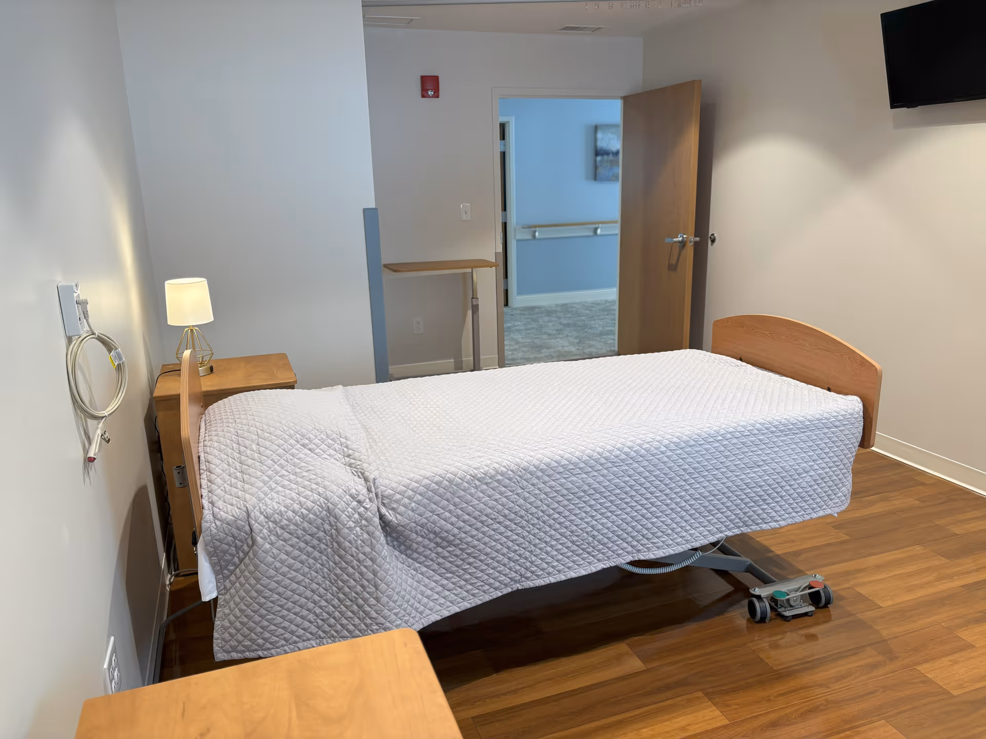 A simple senior living bedroom with an adjustable bed covered in a white quilt, a wooden nightstand and lamp, and a wall-mounted TV.