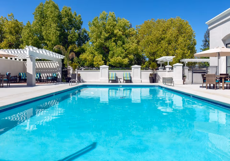 Outdoor swimming pool with clear blue water surrounded by a concrete deck, lounge chairs with striped cushions, tables with umbrellas, and white pergolas. Green trees are visible in the background under a clear blue sky.