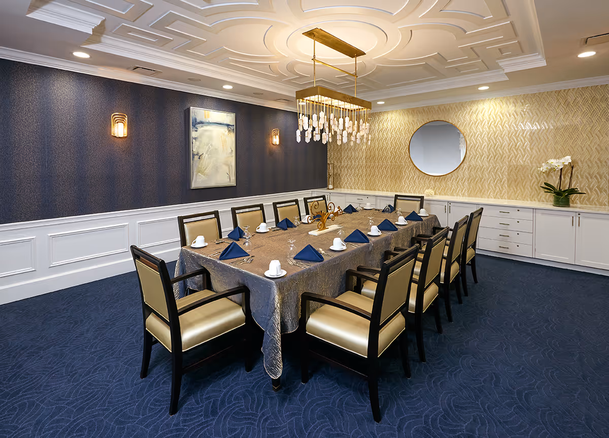 A formal dining room with a long rectangular table covered with a silver tablecloth, set with white cups, glasses, and blue napkins. The room features beige cushioned chairs with dark wooden frames, a decorative ceiling with recessed lighting, a modern chandelier, a large round mirror on a patterned wall, and a piece of abstract art on a dark blue wall.