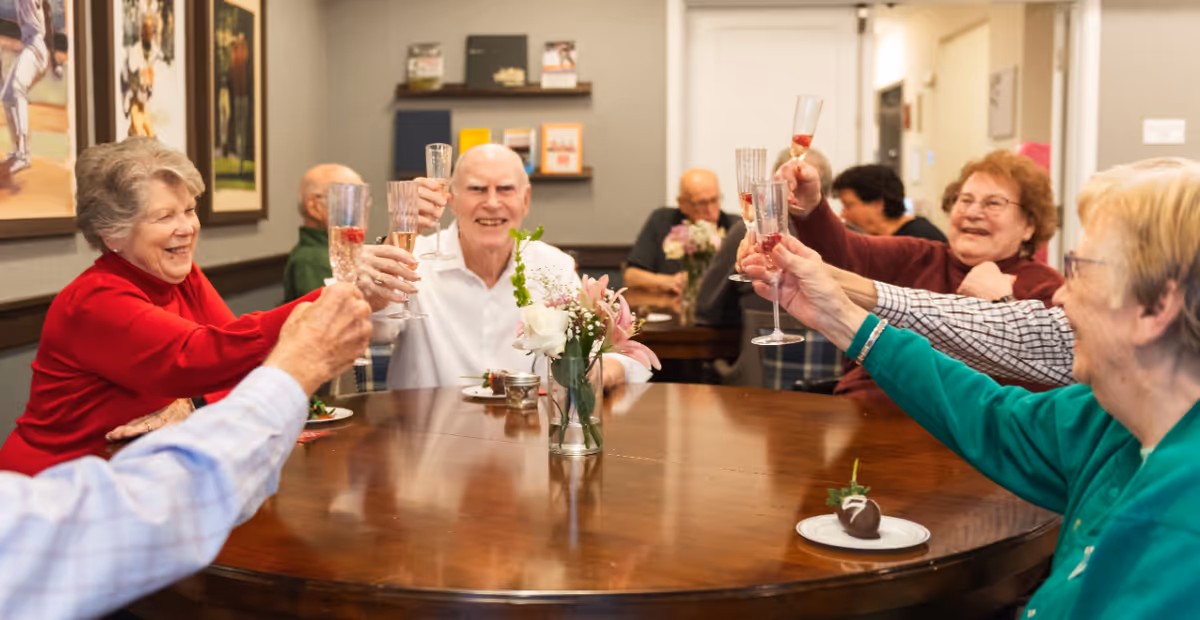A group of elderly people sitting around a large wooden table in a senior living facility, raising their glasses in a toast and smiling. The table has a small vase with flowers and some plates with desserts. The room has framed pictures on the wall and a cozy, welcoming atmosphere.