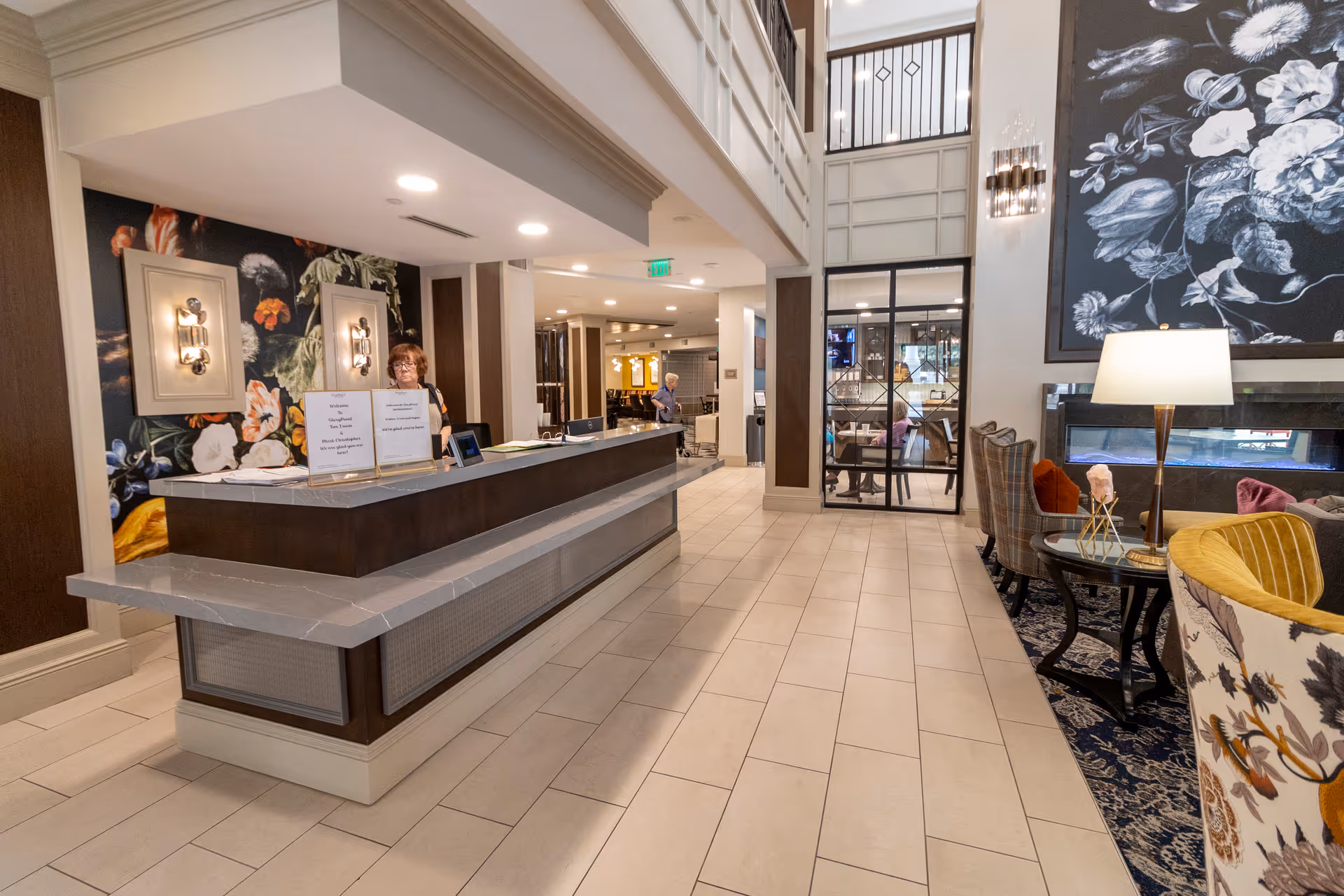 A spacious and well-lit reception area in a senior living facility with a front desk attended by a staff member. The area features modern decor with floral wall art, comfortable seating with patterned chairs, a floor lamp, and a large black and white floral painting above a fireplace. The floor is tiled, and there is a glass-walled room visible in the background.