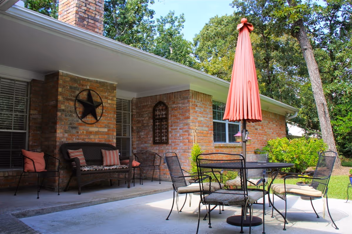 Brick cottage patio with a round table, metal chairs, a red umbrella and wicker bench under a covered porch.