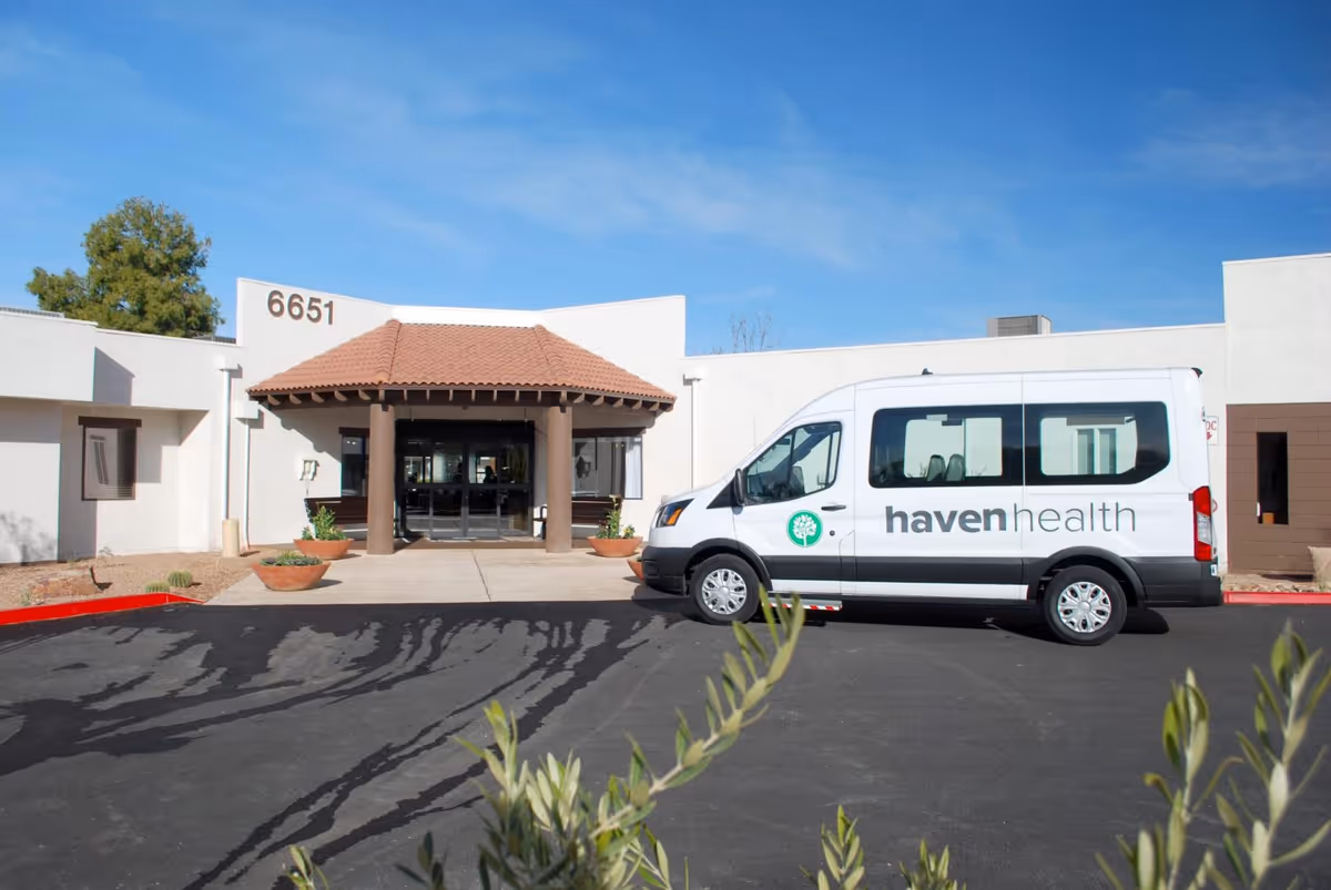 Exterior view of Haven Health Saguaro Valley facility with a white van parked in front. The building has a white facade with a red-tiled roof over the entrance and the number 6651 displayed on the wall. There are some plants and a clear blue sky in the background.
