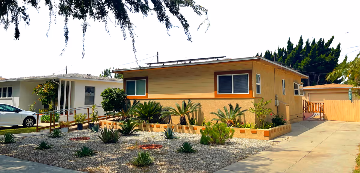 Single-story residential building with beige and yellow exterior walls, surrounded by a landscaped front yard featuring various small plants and shrubs. A concrete driveway leads to a wooden gate on the right side of the building. A white car is parked near the neighboring house on the left. Trees and bushes are visible in the background under a clear sky.