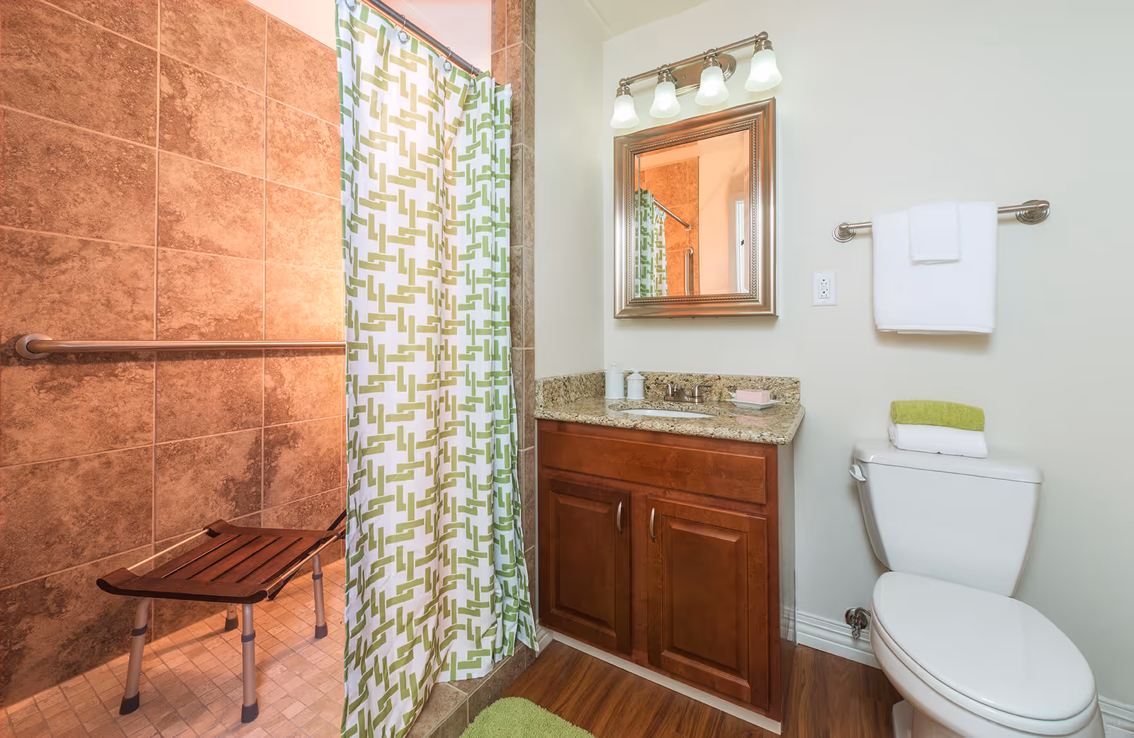Bathroom with a walk-in shower featuring brown tiles and a wooden shower bench, a white toilet with folded towels on top, a wooden vanity with a granite countertop, a mirror with a silver frame, and a towel rack with a white towel. The shower has a curtain with a green and white geometric pattern.
