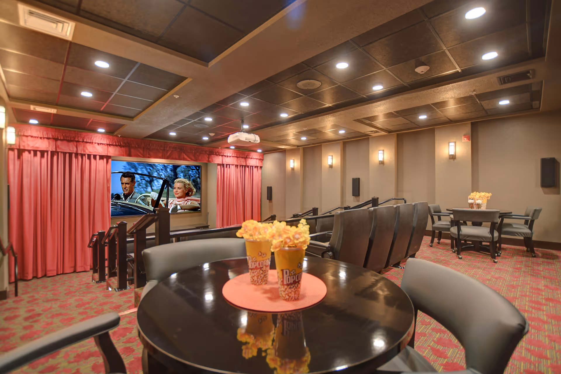 Interior view of a cozy theater room with tiered seating, a large screen showing a classic movie scene, red curtains framing the screen, and round tables with chairs and decorative popcorn containers with flowers on them.